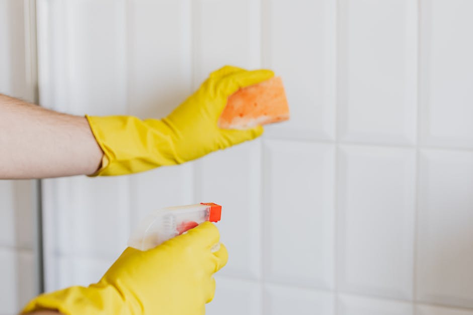 A person wearing yellow rubber gloves is engaged in surface cleaning within a domestic kitchen or bathroom, holding an orange sponge in one hand and a spray bottle in the other. The surface, likely ce