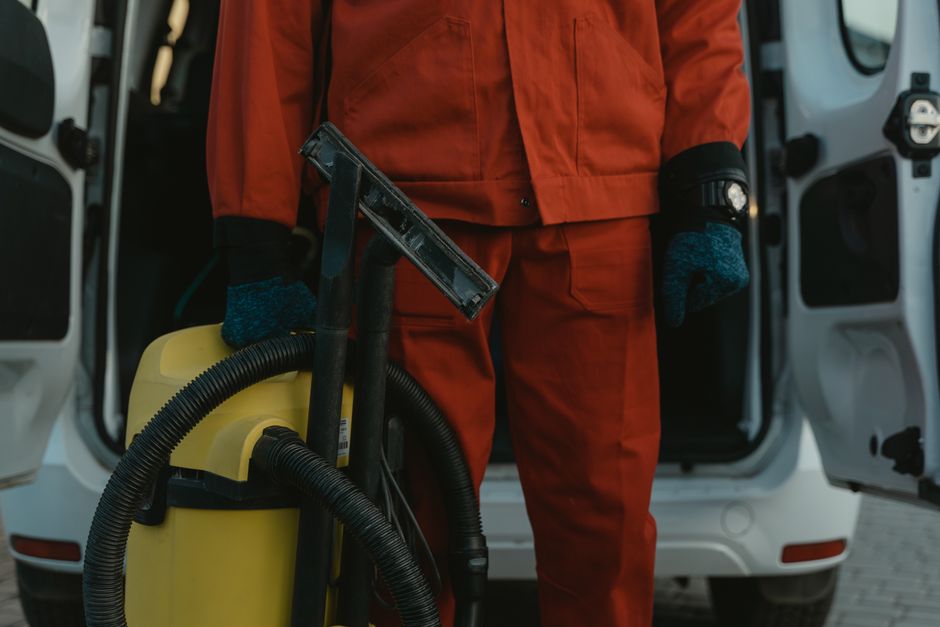 A person wearing yellow rubber gloves is engaged in surface cleaning within a domestic kitchen or bathroom, holding an orange sponge in one hand and a spray bottle in the other. The surface, likely ce